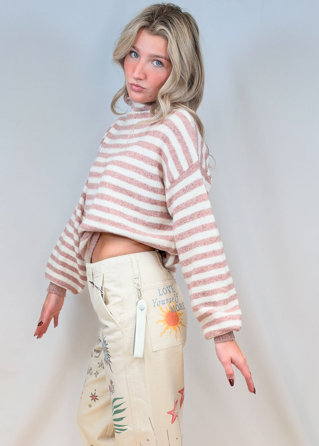 Woman wearing pink and cream stripe sweater with beige doodle pants on plain background