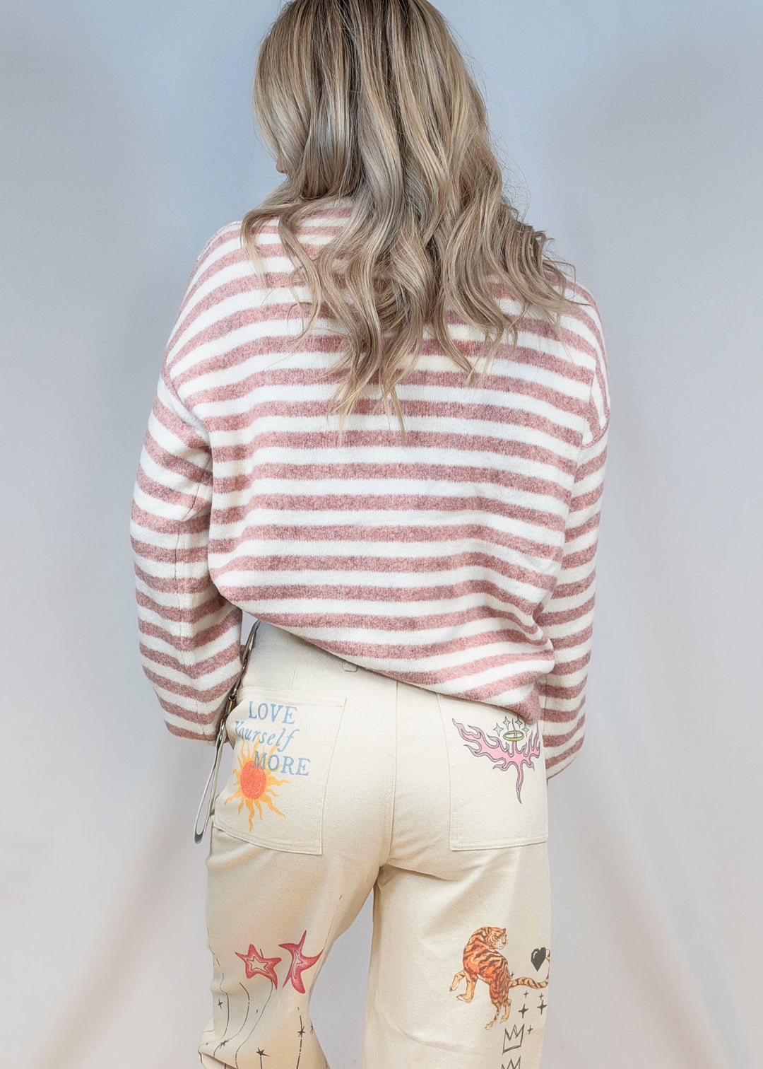 Woman shown from back view wearing pink and cream stripe sweater with beige doodle pants on plain background