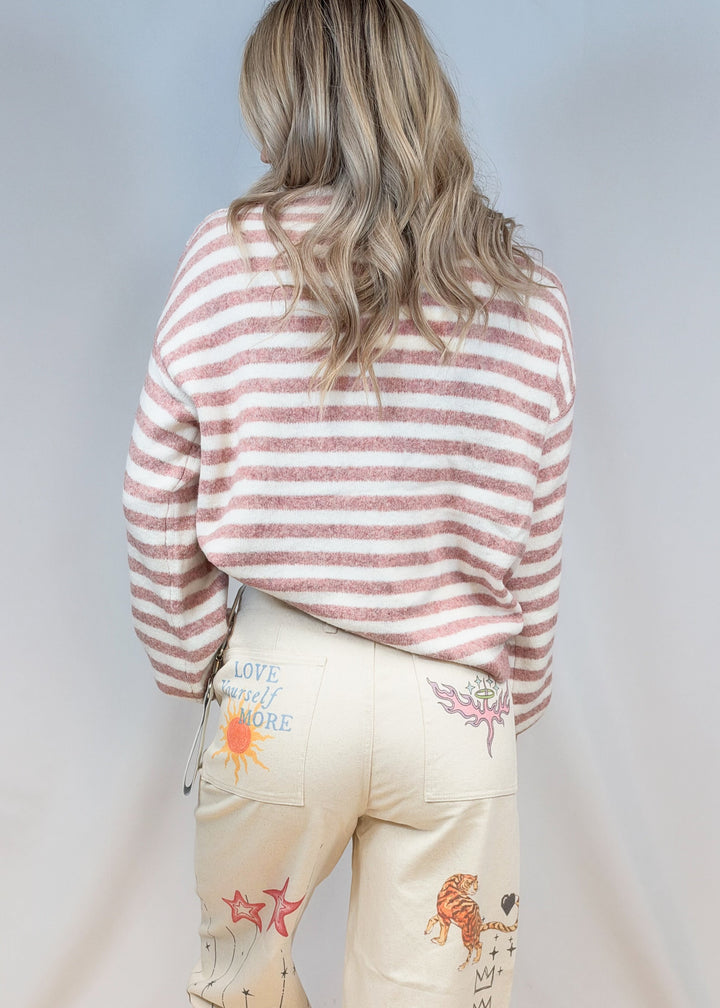Woman shown from back view wearing pink and cream stripe sweater with beige doodle pants on plain background