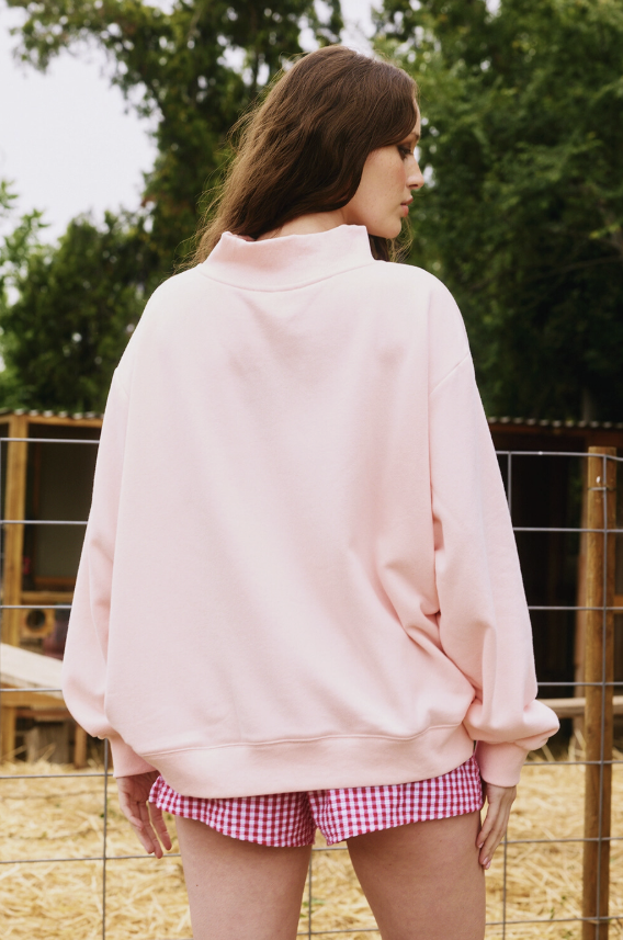 Person wearing a pink sweatshirt and checkered shorts standing outdoors with trees in the background