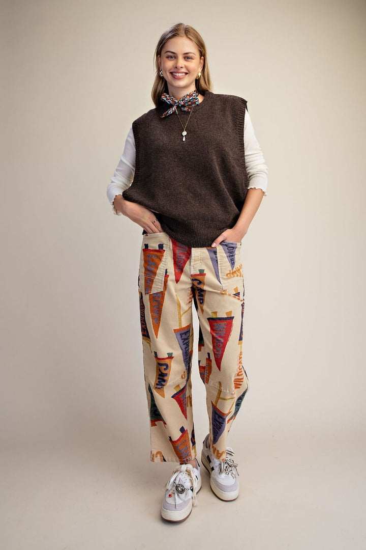 A woman modeling a brown knit sleeveless vest over a white long-sleeve shirt, paired with beige wide-leg pants featuring a colorful geometric pennant pattern, and white sneakers, set against a plain beige studio background.