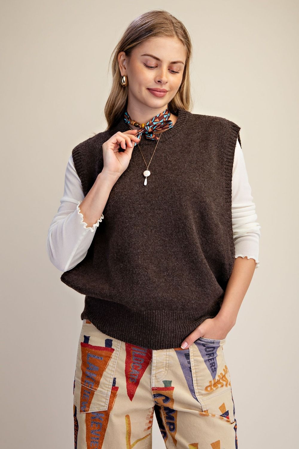 A woman wearing a dark brown sleeveless knit sweater vest over a white long-sleeve shirt and patterned pants, standing against a plain light background.