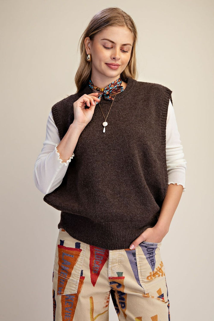 A woman wearing a dark brown sleeveless knit sweater vest over a white long-sleeve shirt and patterned pants, standing against a plain light background.