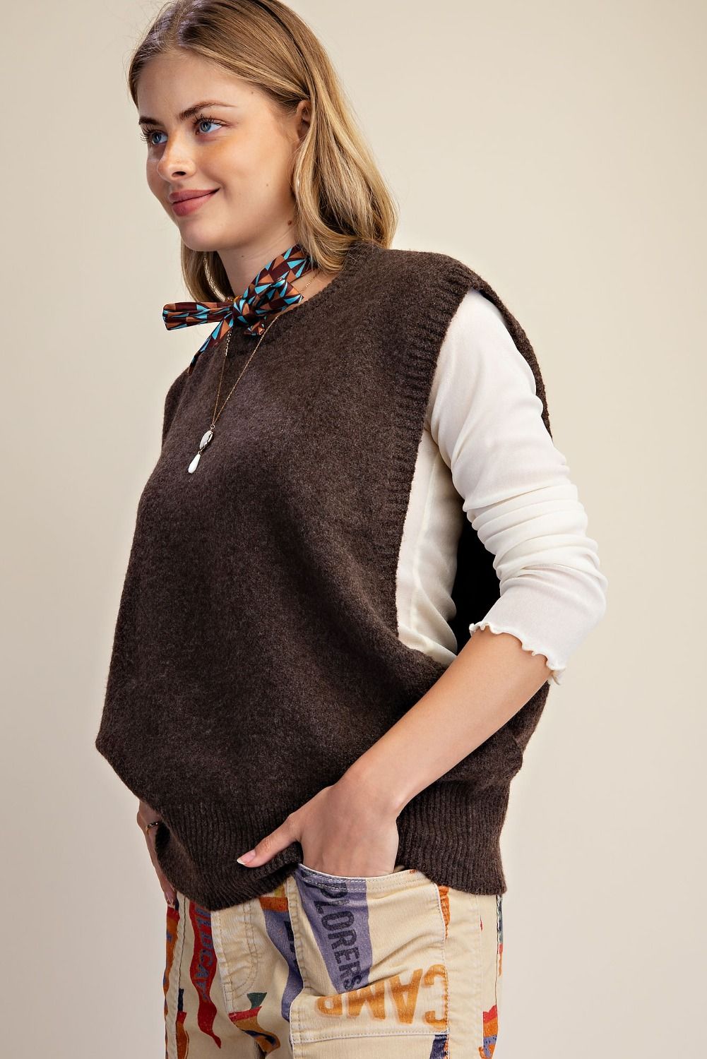A woman wearing a dark brown sleeveless knit vest over a white long-sleeve top and patterned pants, shown from the waist up against a plain light background.
