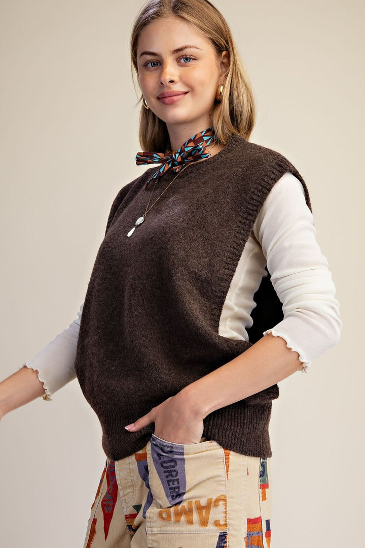A woman wearing a brown knit sweater vest over a white long-sleeve top, accessorized with a patterned neck scarf and a necklace, paired with beige patchwork cargo pants, against a plain beige studio background.