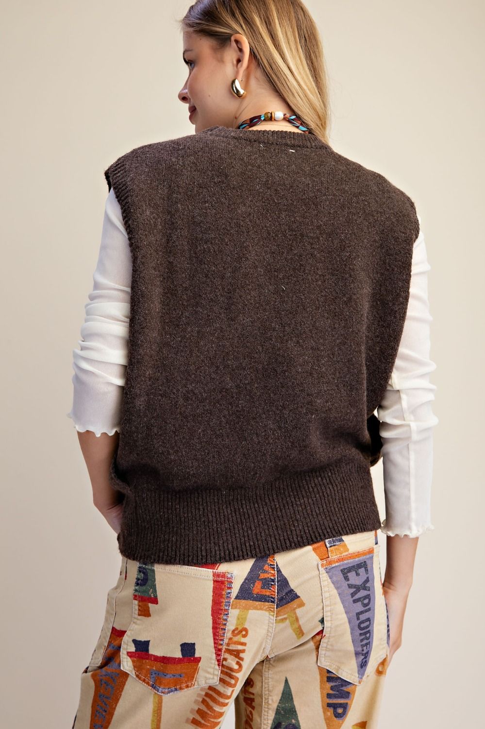 A woman shown from the back wearing a dark brown sleeveless knit sweater vest over a white long-sleeve top and patterned beige pants, against a plain light beige studio background.