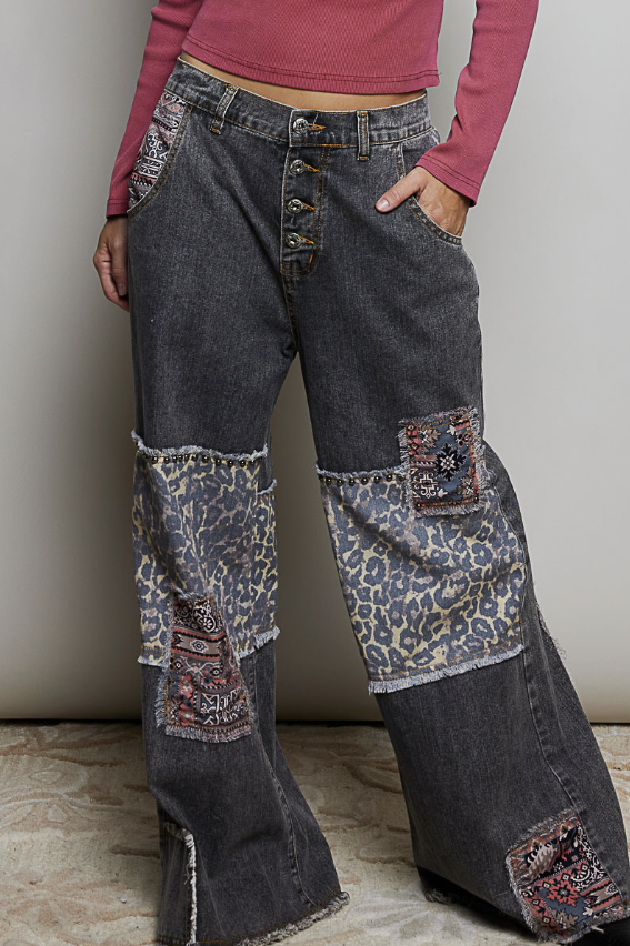 A person wearing dark gray wide-leg patchwork jeans with leopard print and patterned fabric panels on the thighs and lower legs, paired with a pink ribbed long-sleeve top against a plain indoor wall.