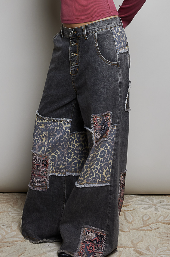 A person wearing wide-leg dark gray patchwork jeans with leopard-print and patterned fabric patches, paired with a pink long-sleeve top, standing against a plain light wall.