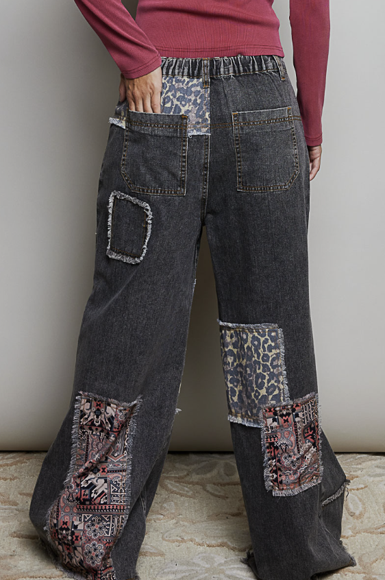 A person wearing dark gray patchwork denim pants with leopard print and floral patterned patches, paired with a dusty rose long-sleeve top, shown from the back against a neutral indoor wall.