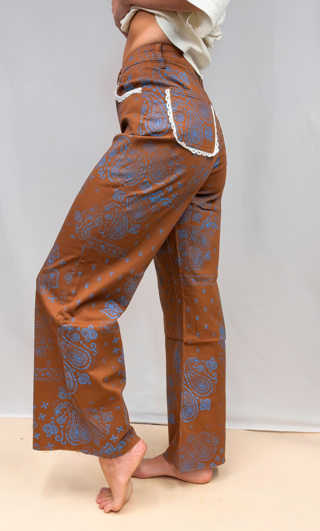 Woman shown from side view wearing brown pants and blue paisley design with lace trim detail on plain background