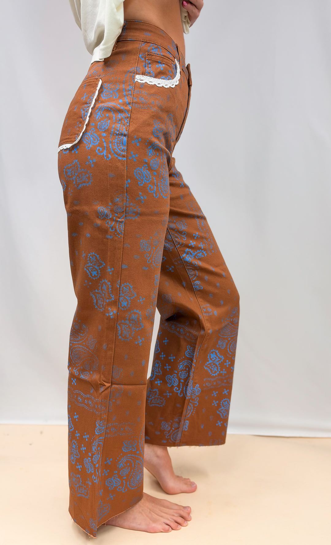 Woman shown from side view wearing brown pants with blue paisley design and lace trim detail on plain background