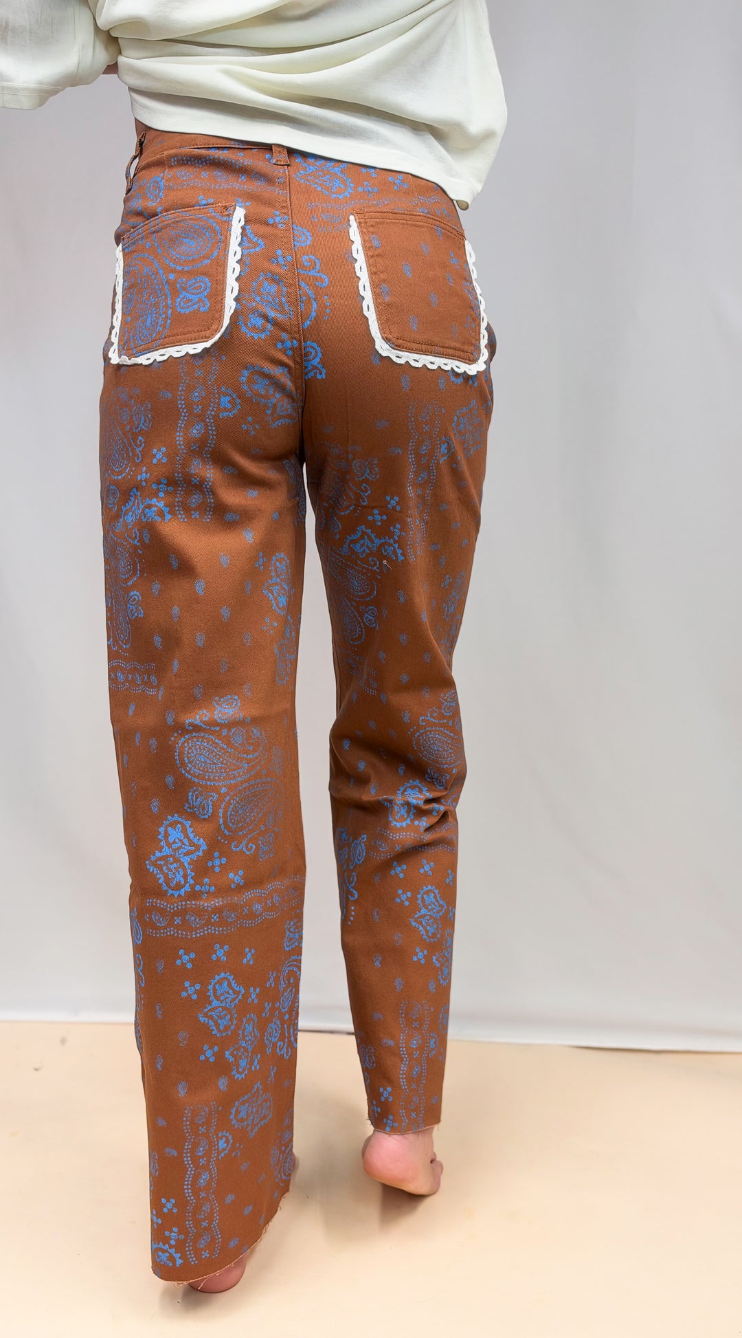 Woman shown from back view wearing brown pants with blue paisley design and lace trim details on plain background
