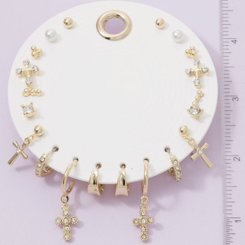 A set of gold-toned hoop and dangle earrings displayed on a white circular stand, with a ruler on the right side for scale. The earrings include cross charms, small hoop pieces, and some pearl accents.