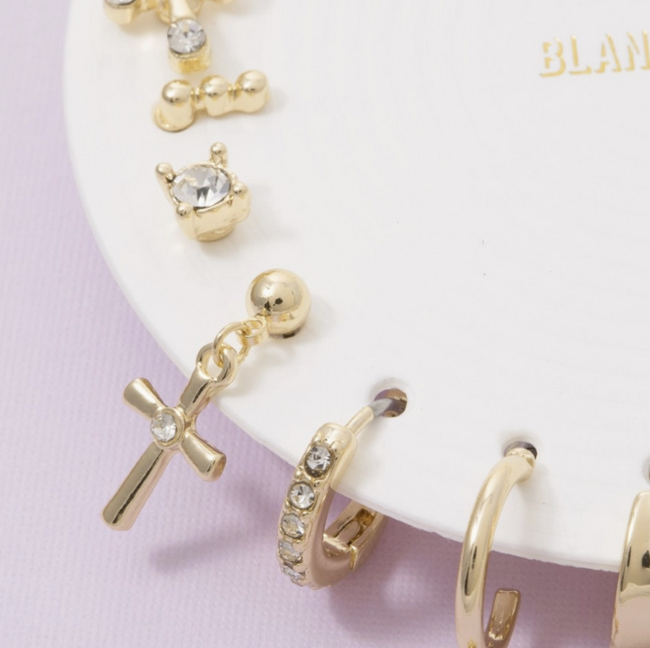 Gold-tone stud earrings displayed on a white circular stand, including a cross charm with a clear rhinestone, a small crown motif, and a huggie hoop with clear rhinestones, against a pale pink background.