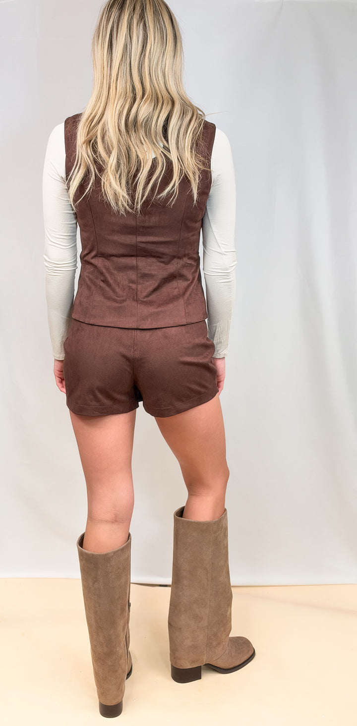 Woman shown from back wearing matching dark brown vest and skort set with knee high boots on plain background