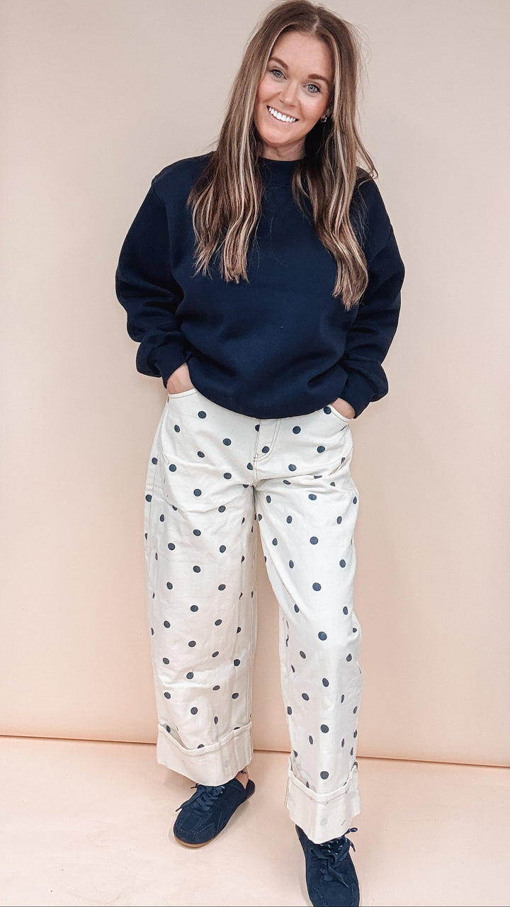 A woman stands in front of a beige backdrop wearing a dark navy sweatshirt, cream wide-leg pants with navy polka dots, and navy shoes.