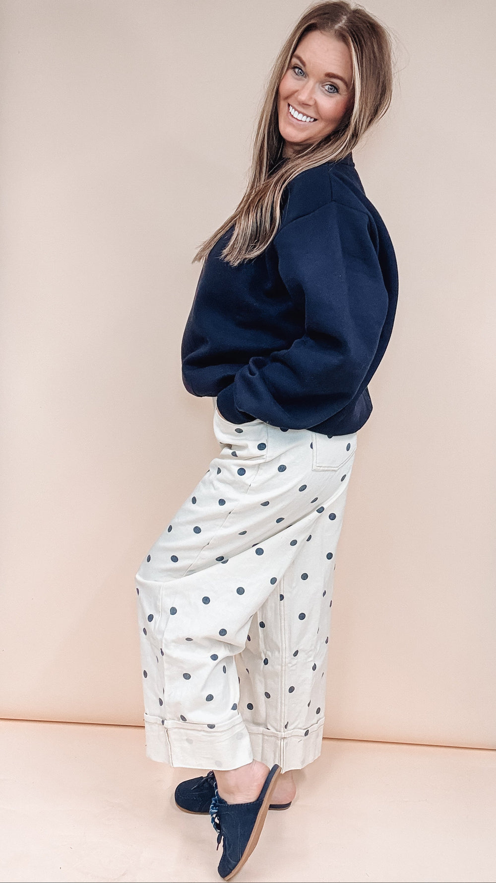 A woman posing in a studio setting wearing a navy blue sweatshirt, white wide-leg pants with navy polka dots, and navy blue slip-on shoes, set against a large beige backdrop.