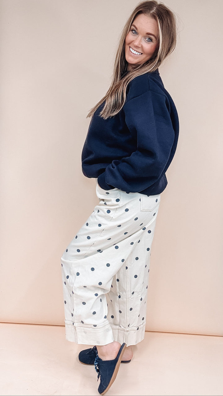 A woman posing in a studio setting wearing a navy blue sweatshirt, white wide-leg pants with navy polka dots, and navy blue slip-on shoes, set against a large beige backdrop.