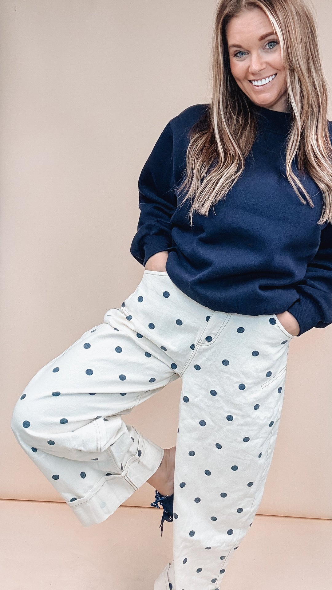 A woman posing in a studio setting wearing a navy blue sweatshirt and white cropped pants with navy blue polka dots.