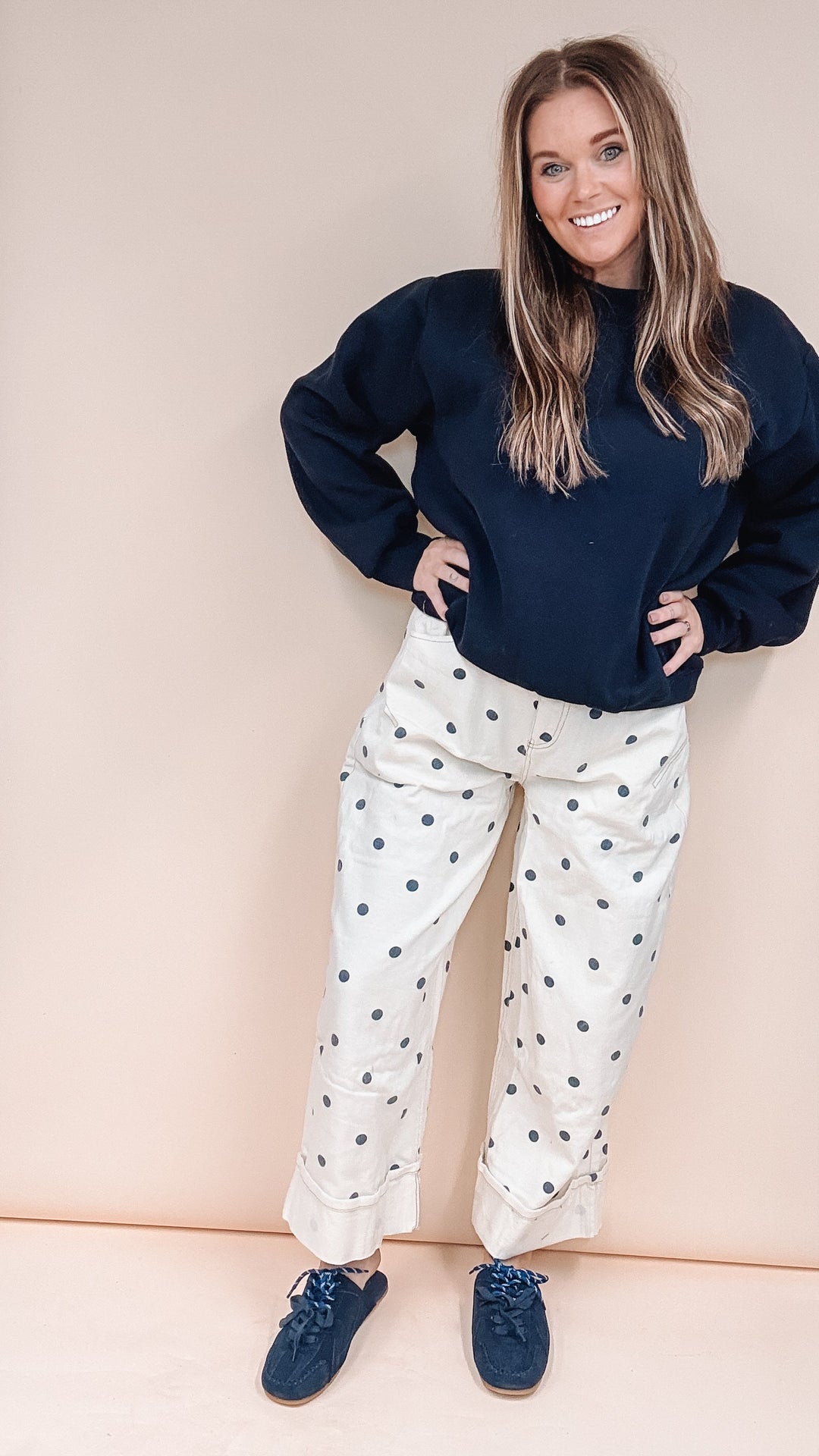 A woman posing in a navy blue sweatshirt and cream pants with dark blue polka dots, standing in front of a beige backdrop in what appears to be a studio or closet space.