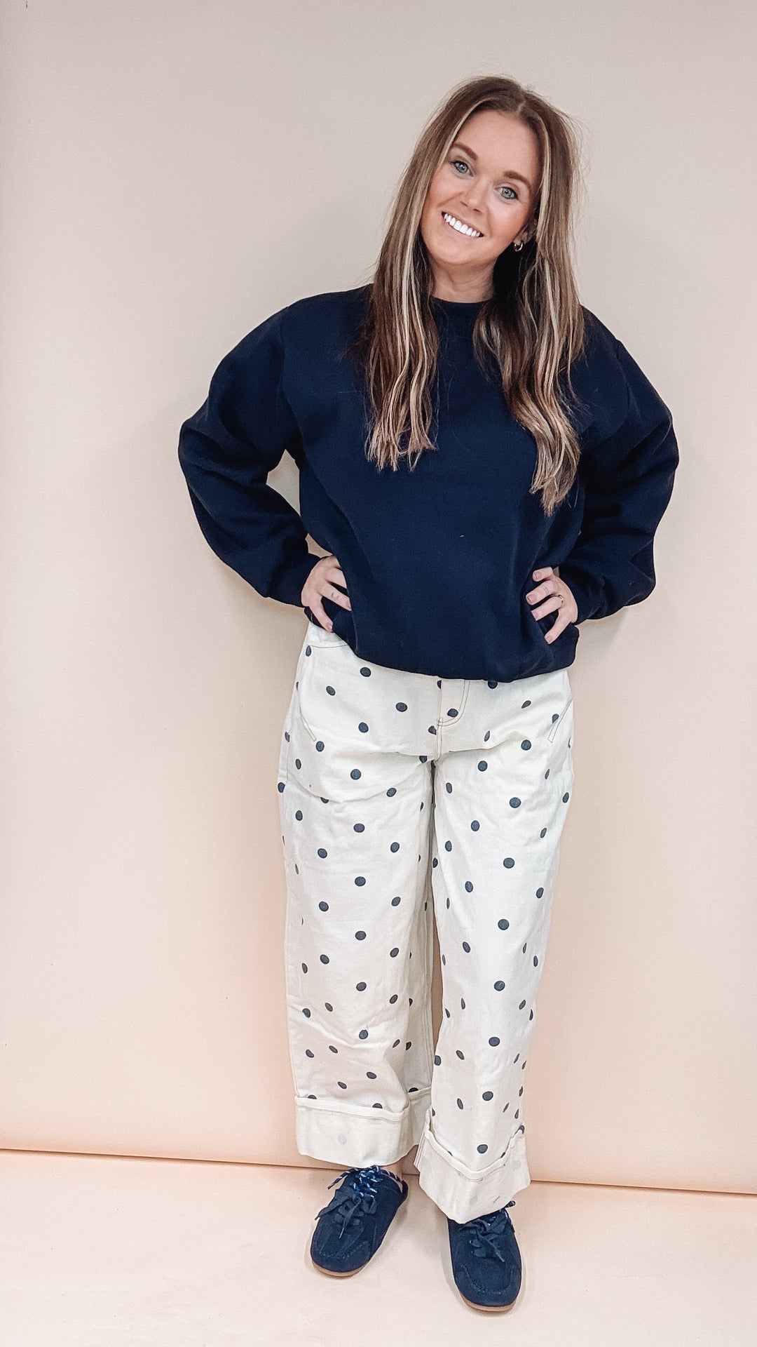A woman stands in front of a beige backdrop wearing a dark navy sweatshirt, cream pants with black polka dots, and navy shoes.