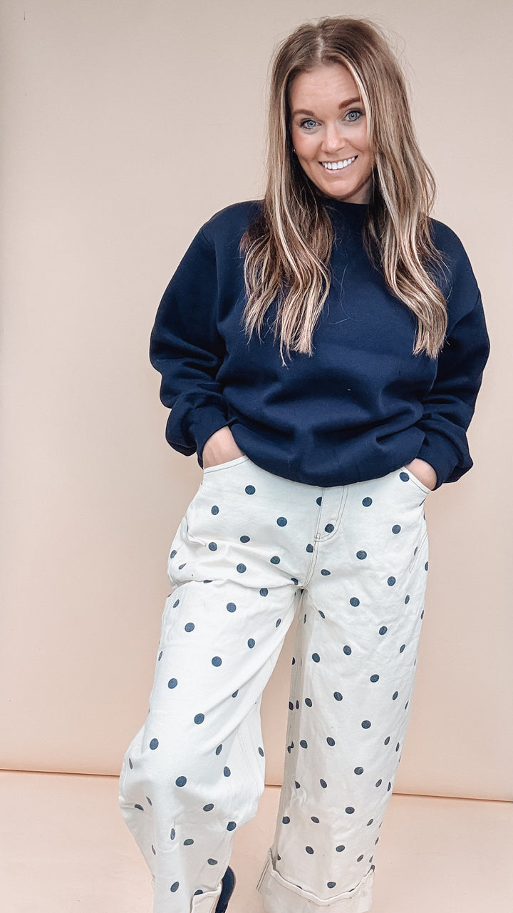 A woman modeling a navy blue mock-neck sweatshirt and white wide-leg trousers with navy blue polka dots, standing in front of a beige backdrop.