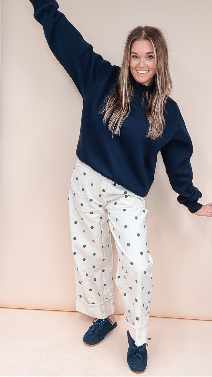 A woman posing in a navy blue sweatshirt and cream wide-leg pants with dark blue polka dots, standing in front of a beige studio backdrop.