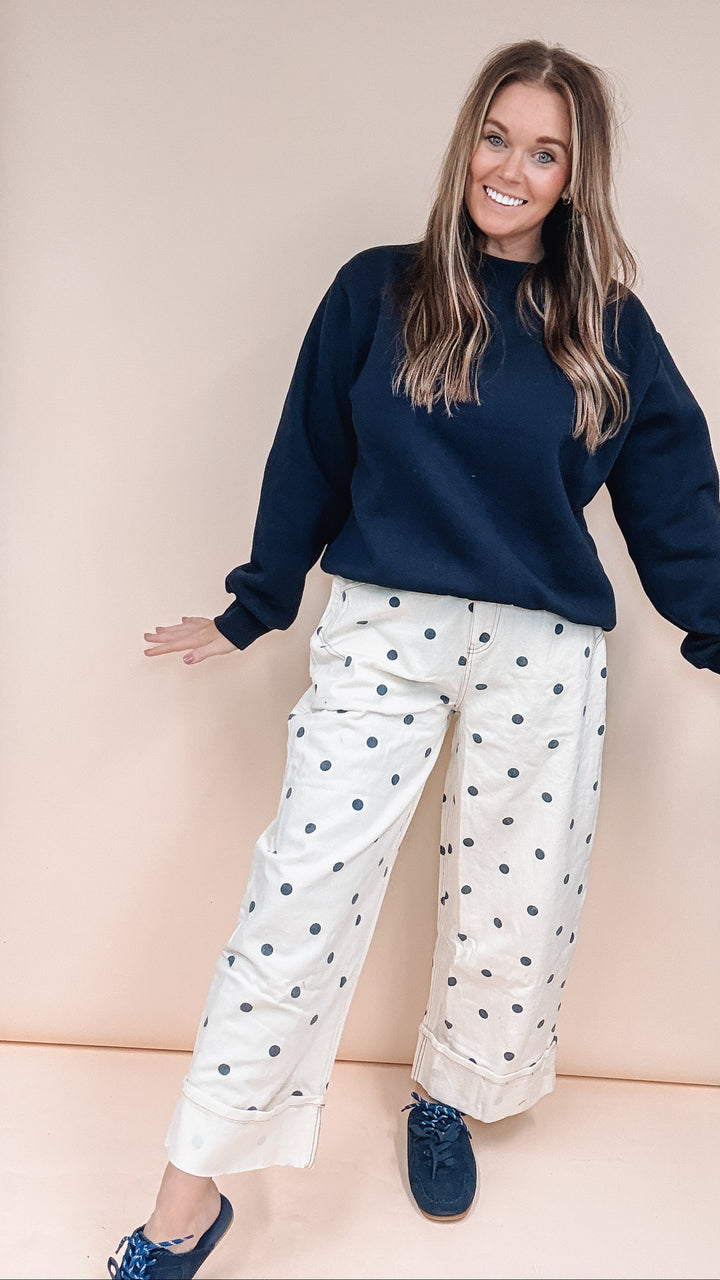 A woman posing in a navy blue sweatshirt and white wide-leg pants with dark blue polka dots, standing in front of a beige studio backdrop.