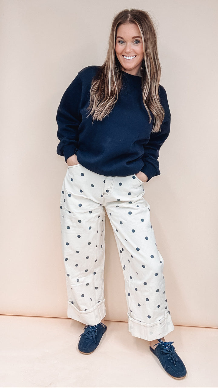 A woman stands in front of a beige backdrop wearing a navy crewneck sweatshirt, white wide-leg pants with dark blue polka dots, and navy slip-on shoes.