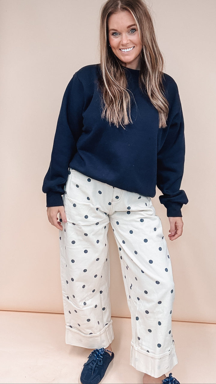 A woman modeling a navy blue crewneck sweatshirt and white wide-leg pants with black polka dots, standing in front of a beige studio backdrop.
