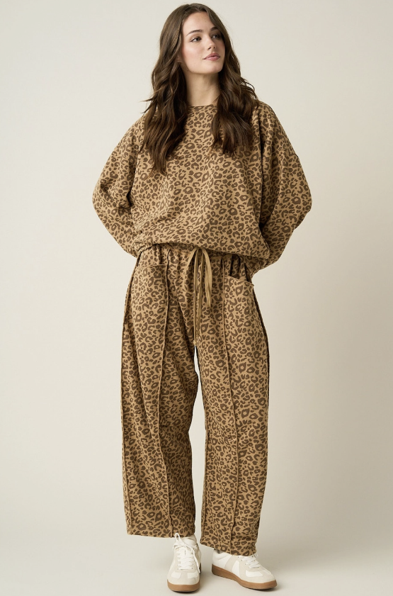 A woman modeling a matching brown and beige leopard-print lounge set, featuring a crewneck sweatshirt and wide-leg drawstring pants, paired with white sneakers against a solid light beige background.