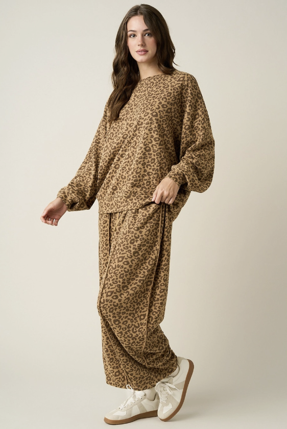 A woman modeling a matching two-piece lounge set with a brown and beige leopard print pattern, consisting of a long-sleeve top and wide-leg pants, paired with white sneakers against a light beige studio background.