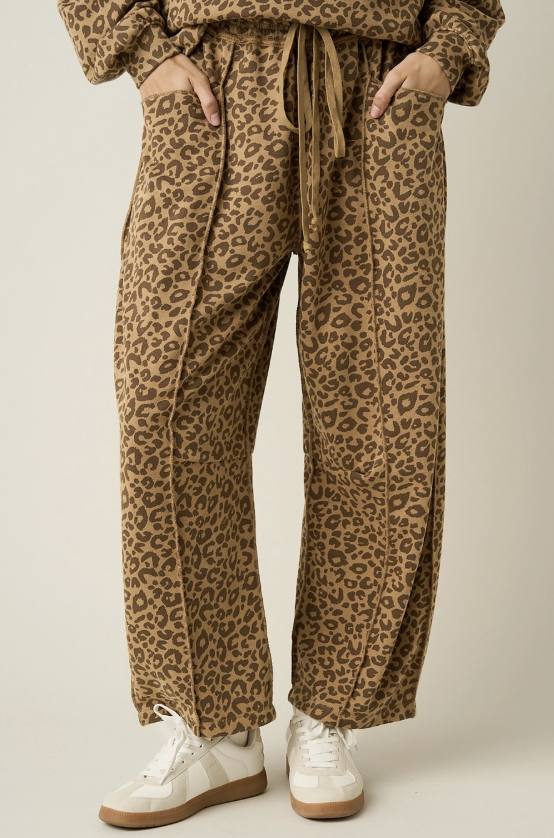 A person wearing loose, wide-leg leopard-print pants with a drawstring waist, shown from the waist down against a plain light beige studio background. The pants have a tan and brown animal pattern, and the model is also wearing white sneakers.