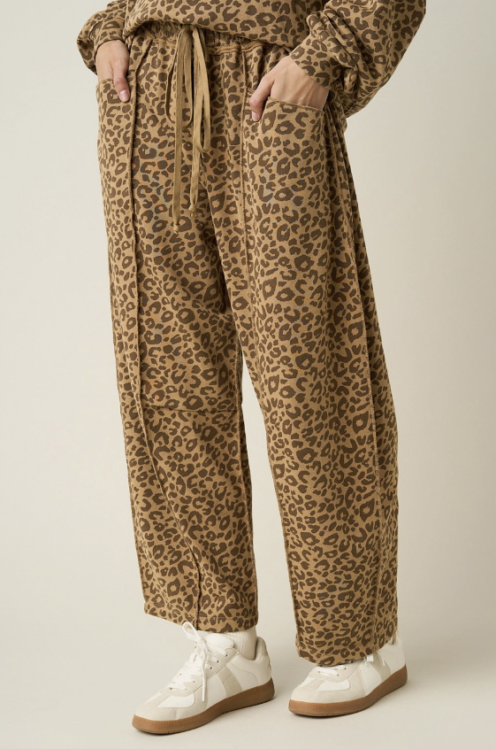 A person wearing loose, wide-leg leopard-print pants with a drawstring waist, shown from the waist down against a plain light beige studio background. The pants have a tan and brown animal pattern with dark brown spots, paired with white sneakers.