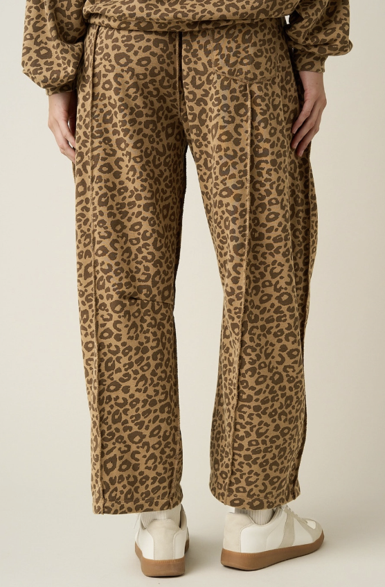 A person shown from the back wearing loose leopard-print pants in tan and brown, with white sneakers, against a plain light beige studio background.