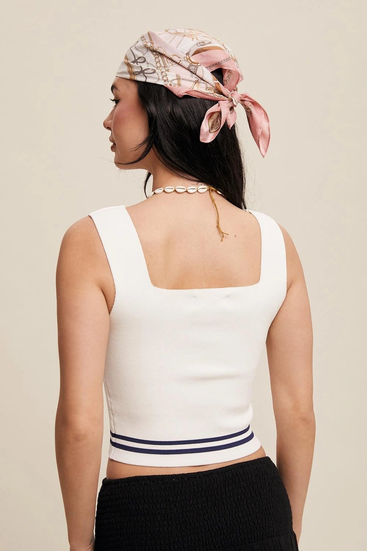 A woman shown from the back wearing a white knit crop top with navy stripes at the hem, a pink patterned headscarf, and a pearl necklace against a light beige studio background.
