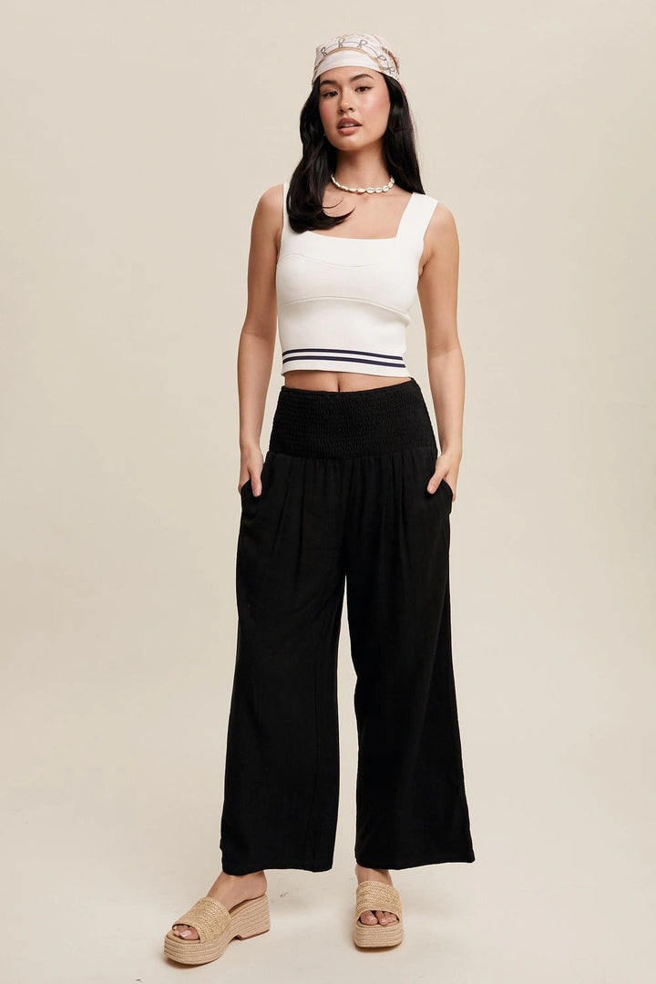 A woman modeling a white ribbed knit crop top with navy stripes, paired with high-waisted black wide-leg pants and beige platform sandals, against a solid beige studio background.