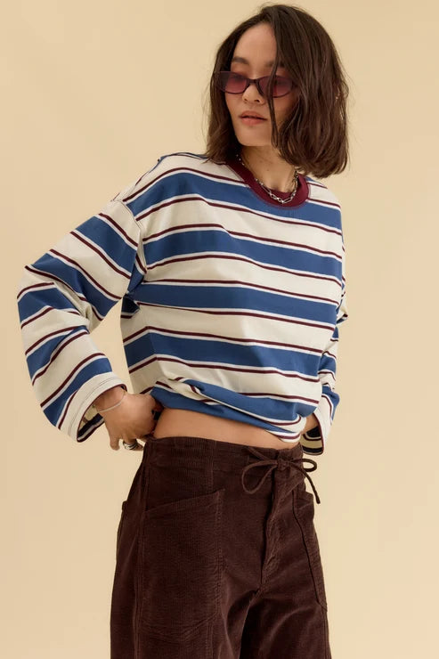 A woman wearing a cropped long-sleeve striped top with blue, white, and thin red stripes, paired with dark brown corduroy pants, against a solid beige background.