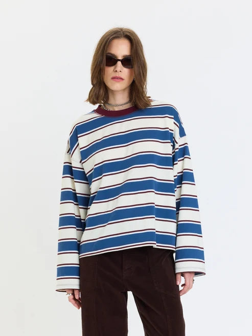 A woman wearing a blue, white, and dark red striped long-sleeve knit top with dark brown pants, photographed against a plain white studio background.