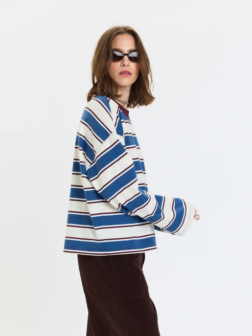 A woman wearing a blue, white, and navy striped long-sleeve knit top with a high collar, paired with dark brown pants and black sunglasses, photographed against a solid white studio background.