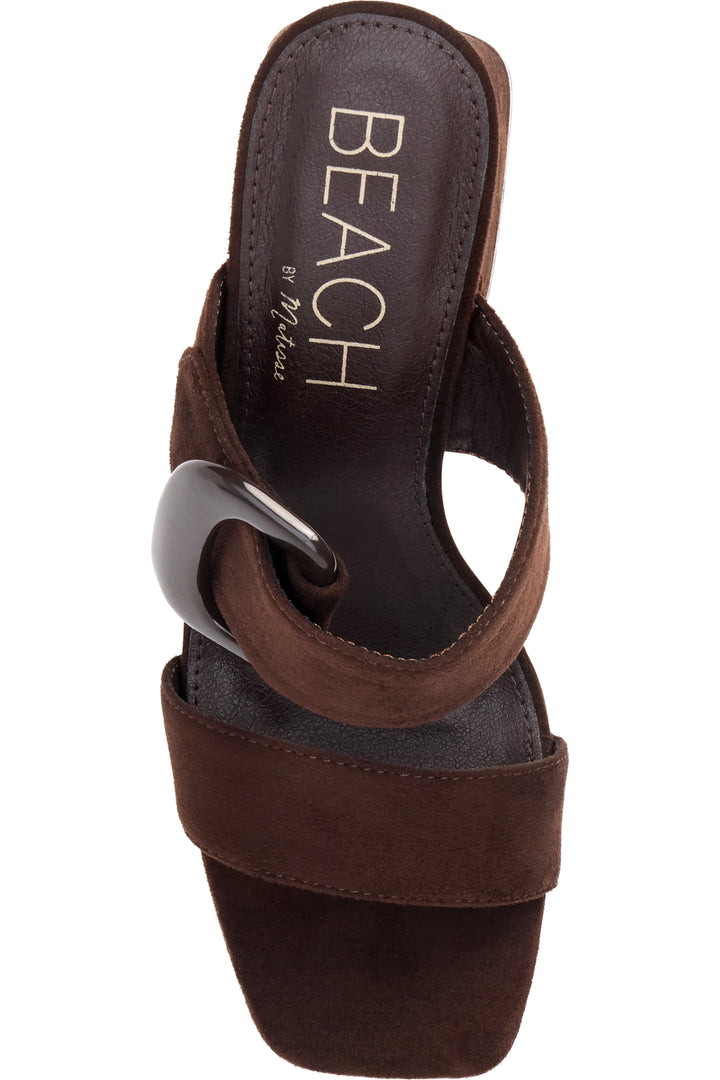 A single dark brown suede slide sandal with a wide strap featuring a large, glossy black ring detail, shown from a top-down view on a white background. The insole displays the brand name &