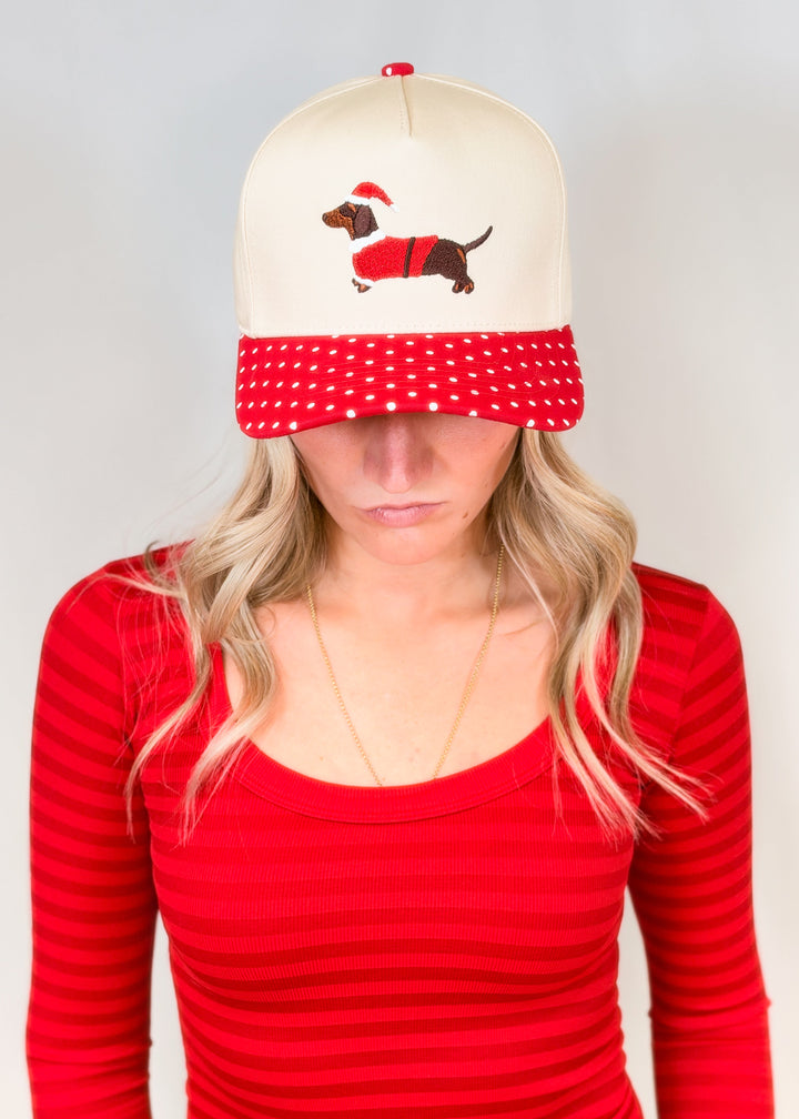 Woman wearing cream color hat with red polka dot brim and embroidered doxie wearing santa coat and hat on plain background