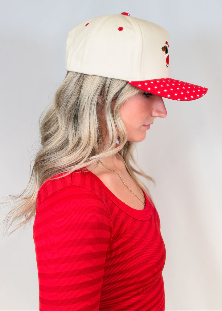 Woman shown from side view wearing cream color hat with red polka dot brim and embroidered doxie on front with red shirt on plain background