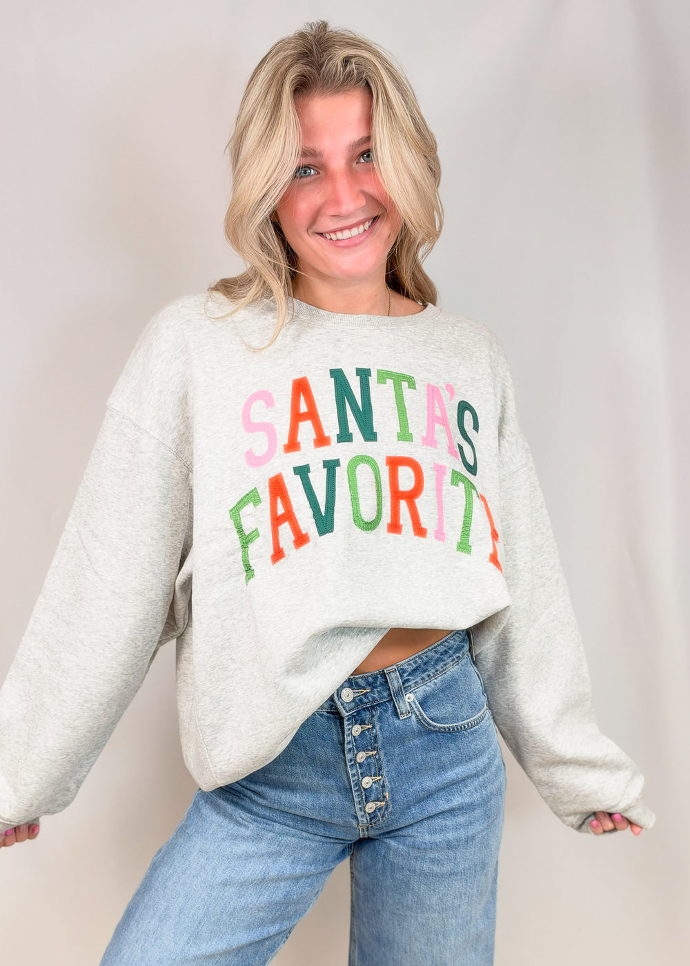 Woman wearing grey sweatshirt with 'SANTAS FAVORITE' on front with jeans on plain background
