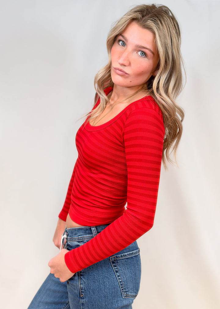 Woman shown from side view wearing red body fitting long sleeve top with jeans on plain background