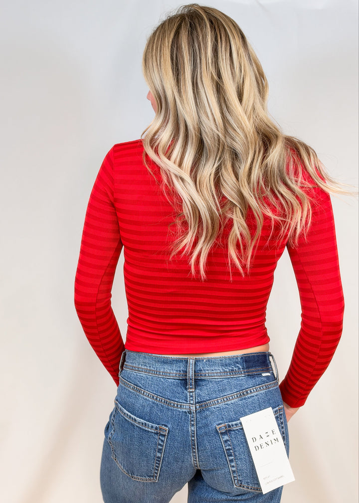 Woman shown from back view wearing body fitting red striped long sleeve top with jeans on plain background