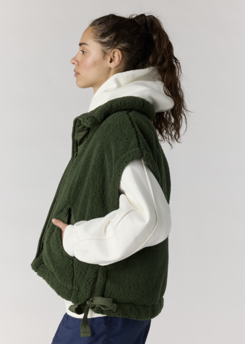 A woman shown in side profile wearing a dark green sherpa fleece hooded vest over a white hoodie, paired with navy pants, against a plain light gray studio background.