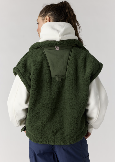 A woman shown from the back wearing a dark green sherpa fleece hooded vest over a white hoodie and navy pants, photographed against a light gray studio background.