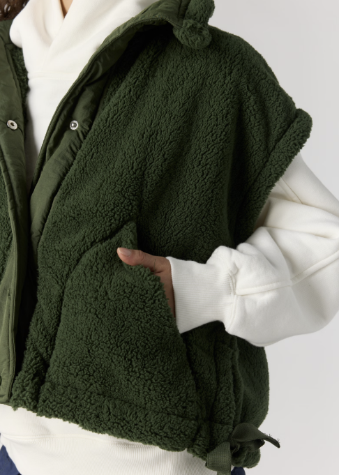 An olive green sherpa fleece hooded vest worn over a white sweatshirt, featuring a drawstring hem and side pockets.