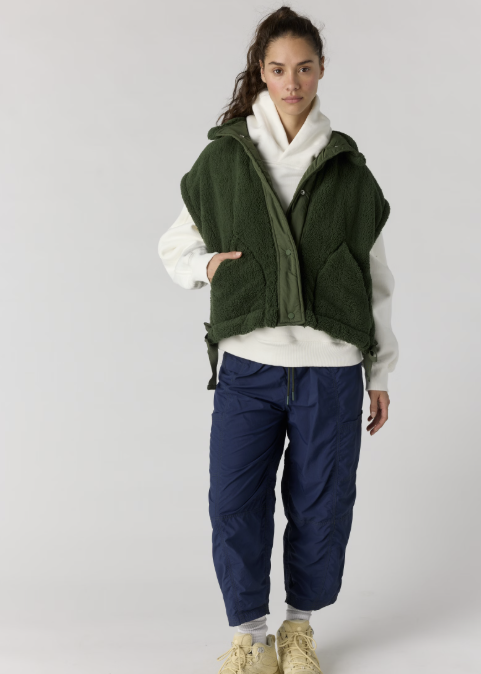 Woman wearing dark green sherpa vest over white hoodie with blue pants and yellow hiking boots on plain background